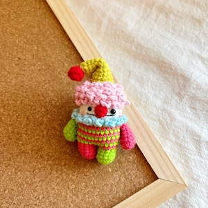 May include: A small crocheted clown doll with a pink and white hat, green and pink striped shirt, and red nose.