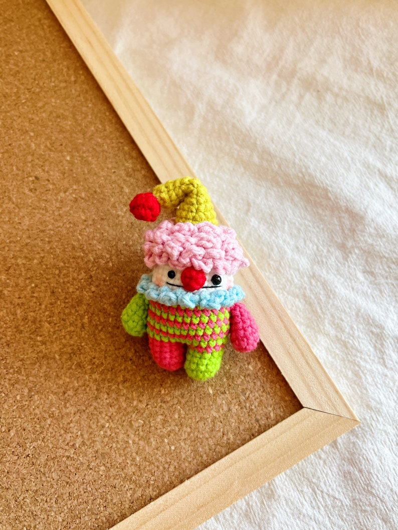 May include: A small crocheted clown doll with a pink and white hat, green and pink striped shirt, and red nose.