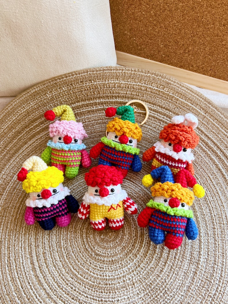 May include: A set of six colorful crocheted clown keychains. Each keychain features a different color scheme and a unique design. The keychains are made with a variety of yarn colors, including red, yellow, blue, green, pink, and white.