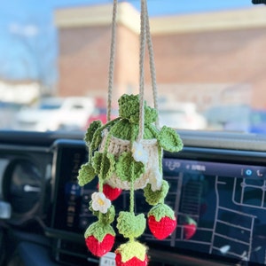 May include: A crocheted strawberry hanging car air freshener. The strawberry is red with green leaves and white flowers. The air freshener is hanging from a white string.