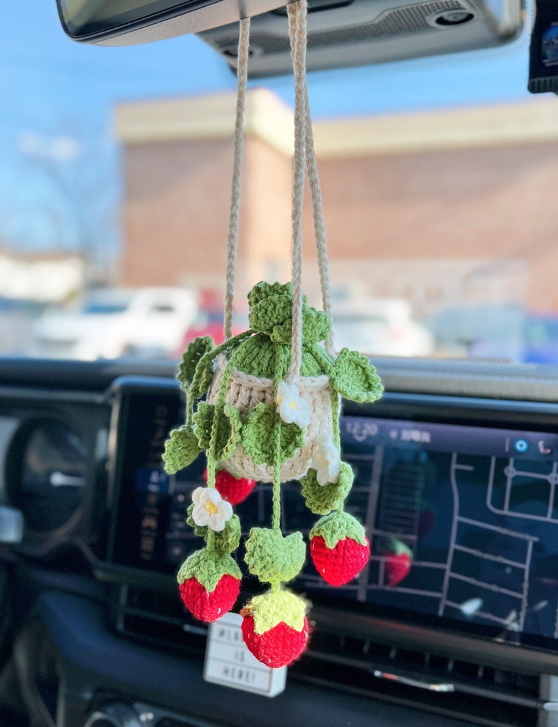 May include: A crocheted strawberry hanging car air freshener. The strawberry is red with green leaves and white flowers. The air freshener is hanging from a white string.