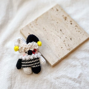 May include: A small crocheted jester doll with a black and white checkered costume, yellow and black hat, and red and white face. The doll is sitting on a light brown textured surface.