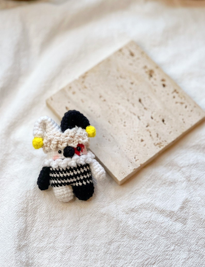 May include: A small crocheted jester doll with a black and white checkered costume, yellow and black hat, and red and white face. The doll is sitting on a light brown textured surface.