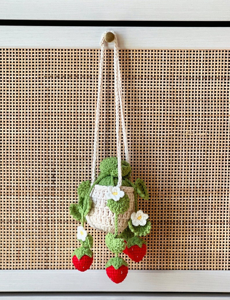 May include: A crocheted strawberry-shaped hanging planter with a cream-colored pouch and green leaves. The planter has three red strawberries hanging from it.