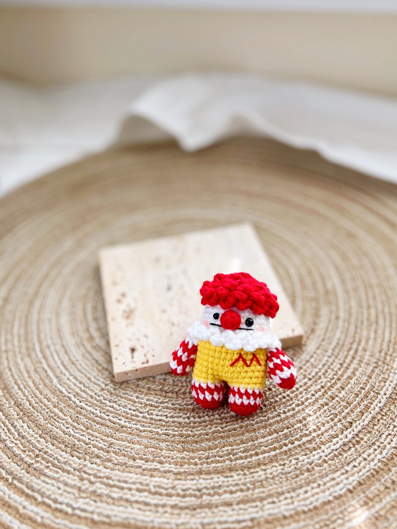 May include: A crocheted clown doll with a red and white striped outfit, yellow body, and red hair. The doll is sitting on a woven straw mat.