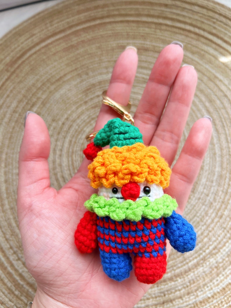 May include: A crocheted clown keychain with a green hat, orange hair, white face, red nose, and blue and red checkered outfit.