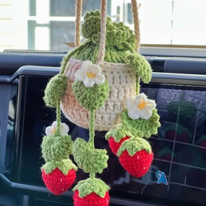 May include: A crocheted strawberry hanging decoration with a white flower and green leaves. The decoration is hanging from a rear view mirror in a car.
