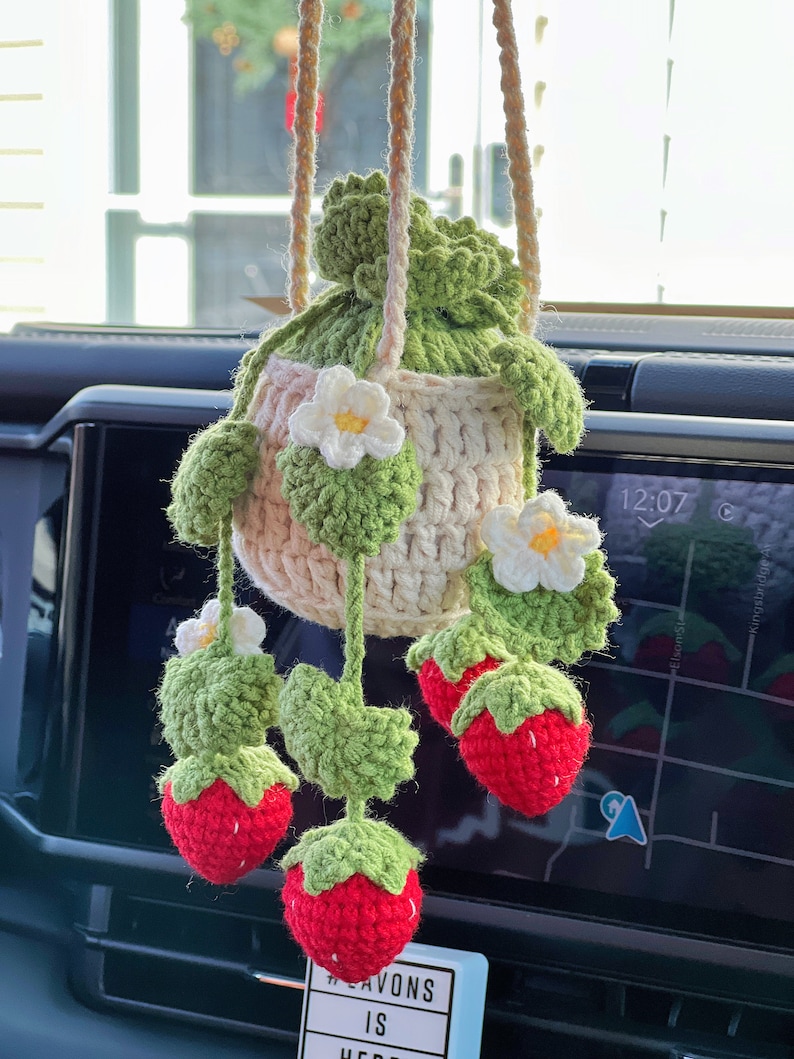 May include: A crocheted strawberry hanging decoration with a white flower and green leaves. The decoration is hanging from a rear view mirror in a car.