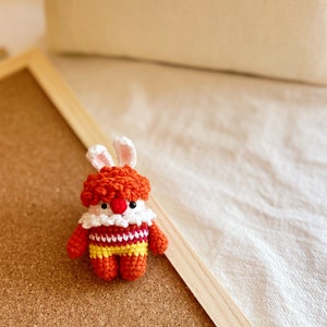 May include: A small crocheted bunny toy with orange and yellow striped clothing and a white collar. The bunny has a red nose and white eyes.