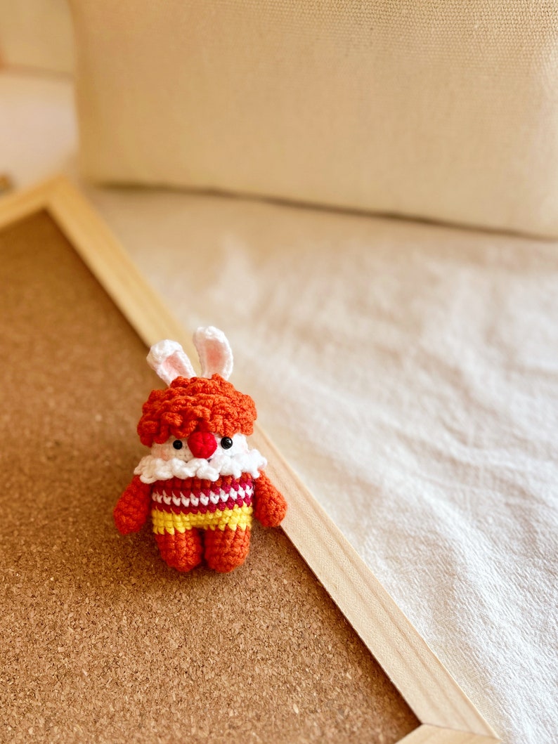 May include: A small crocheted bunny toy with orange and yellow striped clothing and a white collar. The bunny has a red nose and white eyes.