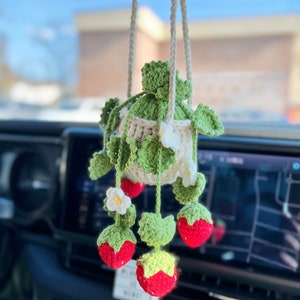 May include: A crocheted strawberry hanging ornament with a white pot and green leaves. The ornament is hanging from a rearview mirror in a car.