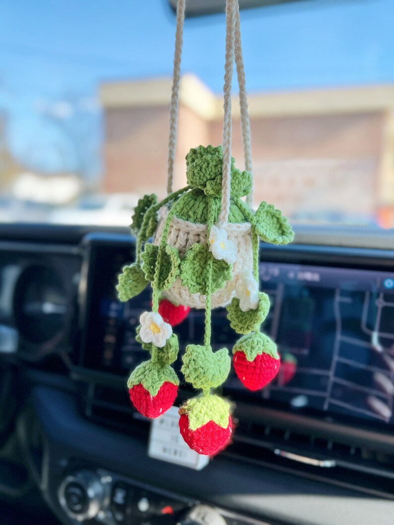 May include: A crocheted strawberry hanging ornament with a white pot and green leaves. The ornament is hanging from a rearview mirror in a car.