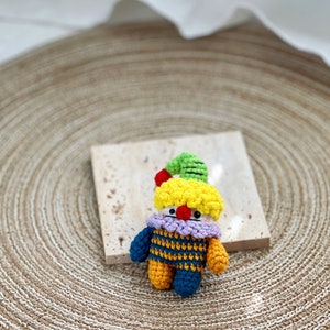 May include: A small crocheted clown doll with a yellow head, green hat, purple and blue striped shirt, and yellow and blue legs.