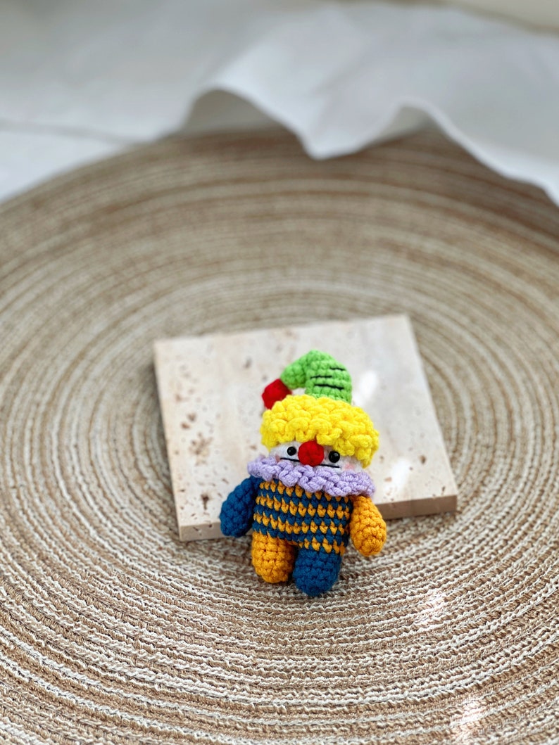May include: A small crocheted clown doll with a yellow head, green hat, purple and blue striped shirt, and yellow and blue legs.