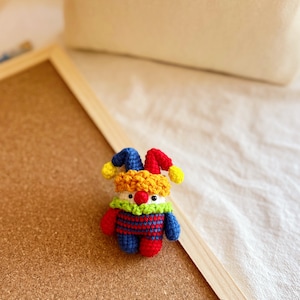 May include: A small crocheted jester doll with a red, yellow, and blue costume. The doll has a white face with a red nose and green eyes.