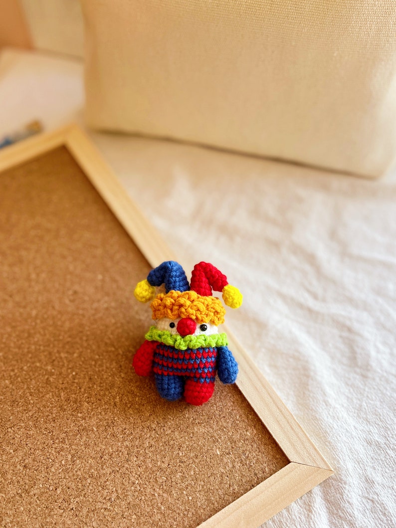 May include: A small crocheted jester doll with a red, yellow, and blue costume. The doll has a white face with a red nose and green eyes.