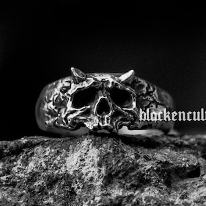 May include: A silver skull ring with horns, detailed with a textured surface. The ring is resting on a rough, grey rock.