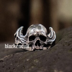 May include: A silver skull ring with intricate details. The skull has a detailed face with a prominent nose and jawline. The ring features two wings on either side of the skull.
