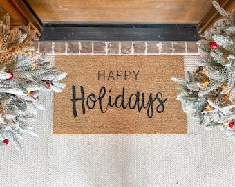 May include: A rectangular coir doormat with the words "HAPPY Holidays" in black script. The mat is placed in front of a door, flanked by two small, snow-covered Christmas trees decorated with red ornaments.