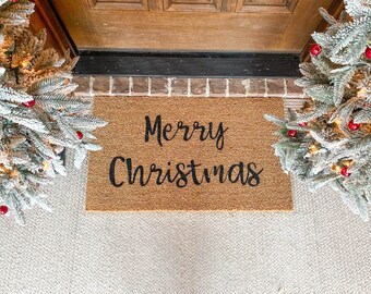 May include: A brown coir doormat with the text "Merry Christmas" in black cursive lettering. The doormat is placed on a white carpet in front of a wooden door.