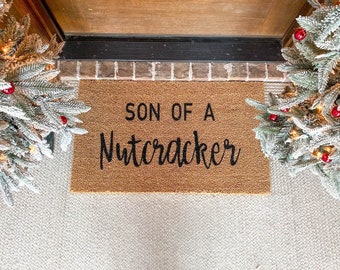 May include: A brown coir doormat with the text "SON OF A NUTCRACKER" in black lettering.