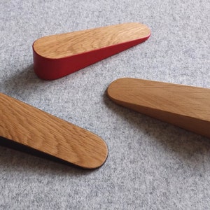 May include: Three wooden doorstops in different colors. The doorstop on the left has a black bottom and a natural wood top. The doorstop in the center has a natural wood finish. The doorstop on the right has a red bottom and a natural wood top.
