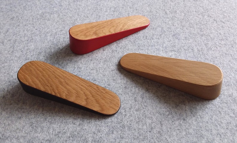May include: Three wooden doorstops in different colors. The doorstop on the left has a black bottom and a natural wood top. The doorstop in the center has a natural wood finish. The doorstop on the right has a red bottom and a natural wood top.