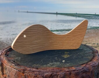 May include: A wooden fish-shaped ornament with a hole in the side, sitting on a weathered wooden stump. The ornament is light brown and has a natural wood grain.