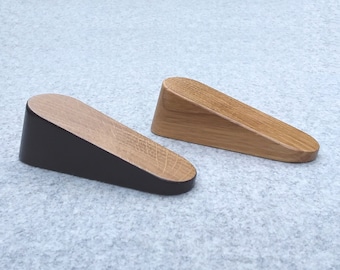 May include: Two wooden doorstops, one with a natural wood finish and one with a black painted finish. Both are wedge-shaped and have a rounded top.