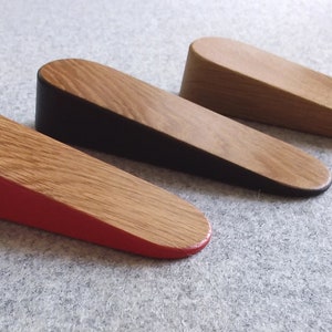 May include: Three wooden door stops in different colours. The door stops are wedge-shaped and have a natural wood finish. One is red, one is black, and one is natural wood.