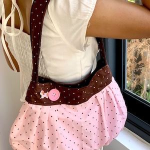 May include: A pink and brown polka dot shoulder bag. The bag features a pink body with small black polka dots and a brown top and strap with white polka dots. A pink button and a small dog-shaped embellishment are on the front.