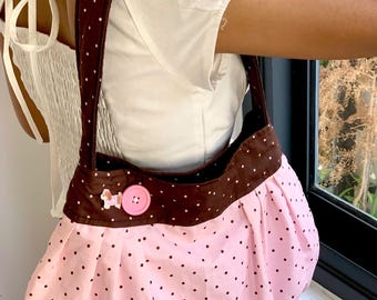May include: A pink and brown polka dot shoulder bag. The bag features a pink body with small black polka dots and a brown top and strap with white polka dots. A pink button and a small dog-shaped embellishment are on the front.