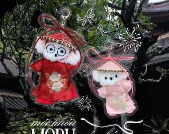 May include: Two plush animal keychains featuring white fur, round glasses, and conical hats. One wears a red outfit with floral embroidery, the other a light pink outfit with similar embroidery. Both include red ribbons and metal key ring attachments.