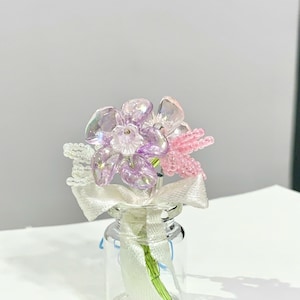 Könnte beinhalten: Eine kleine Glasflasche enthält einen Strauß aus Perlenblumen. Die Blumen sind in Rosa-, Lila- und Klartönen gehalten, mit einem weißen Band um den Flaschenhals gebunden. Die Stiele sind grün.