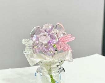 May include: A miniature glass bottle holds a bouquet of beaded flowers. The flowers are in shades of pink, purple, and clear, with a white ribbon tied around the bottle's neck. The stems are green.