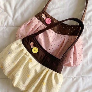 Könnte beinhalten: Zwei kleine, handgefertigte Stoffhandtaschen. Eine ist rosa mit schwarzen Punkten, die andere gelb mit einem kleinen Blumenmuster. Beide haben braune Akzente, Griffe und dekorative Knöpfe.
