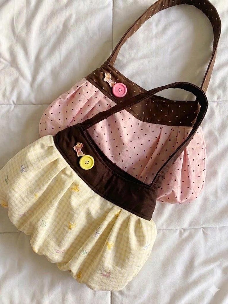 May include: Two small, handmade fabric purses. One is pink with black polka dots, the other is yellow with a small floral pattern. Both have brown accents, handles, and decorative buttons.