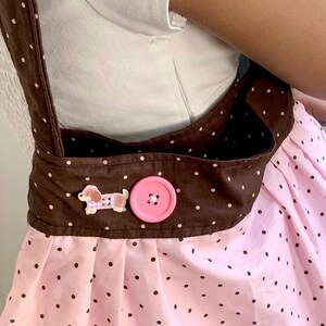 Peut inclure: Un tablier &agrave; pois roses et marron avec une sangle et une poche marron. Le tablier a un bouton rose et une petite d&eacute;coration en forme de chien. Le tablier est port&eacute; sur une chemise blanche.