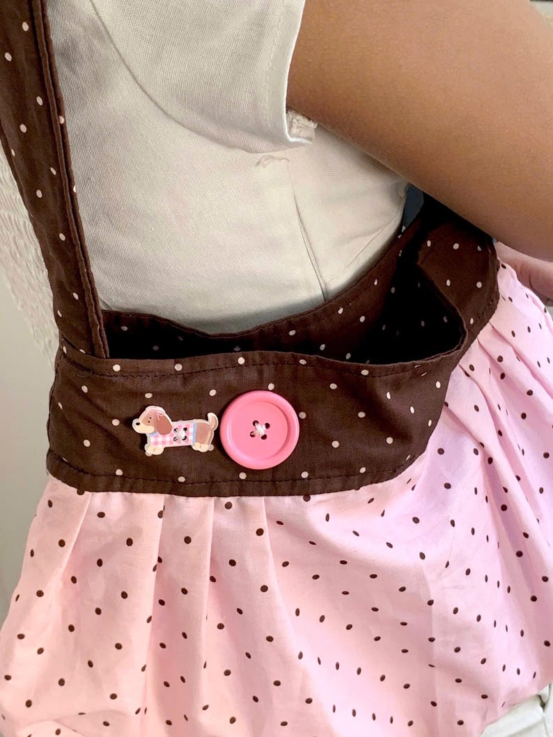 Peut inclure: Un tablier &agrave; pois roses et marron avec une sangle et une poche marron. Le tablier a un bouton rose et une petite d&eacute;coration en forme de chien. Le tablier est port&eacute; sur une chemise blanche.