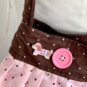 Peut inclure: Tablier rose et marron &agrave; pois avec un bouton en forme de chien et un gros bouton rose. Le tablier a des bretelles marron et une jupe rose &agrave; pois marron. Le bouton en forme de chien porte une tenue rose et bleue.