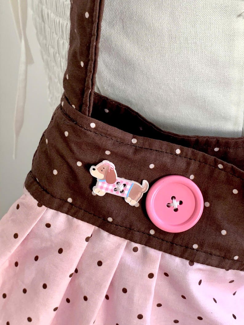 Peut inclure: Tablier rose et marron &agrave; pois avec un bouton en forme de chien et un gros bouton rose. Le tablier a des bretelles marron et une jupe rose &agrave; pois marron. Le bouton en forme de chien porte une tenue rose et bleue.