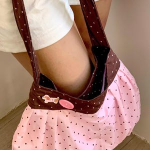 Peut inclure: Un sac &agrave; main rose &agrave; pois avec un dessus et des sangles marron. Le sac est orn&eacute; d'un bouton d&eacute;coratif et d'un petit ornement en forme de chien. Le sac est en tissu et a un design fronc&eacute;.
