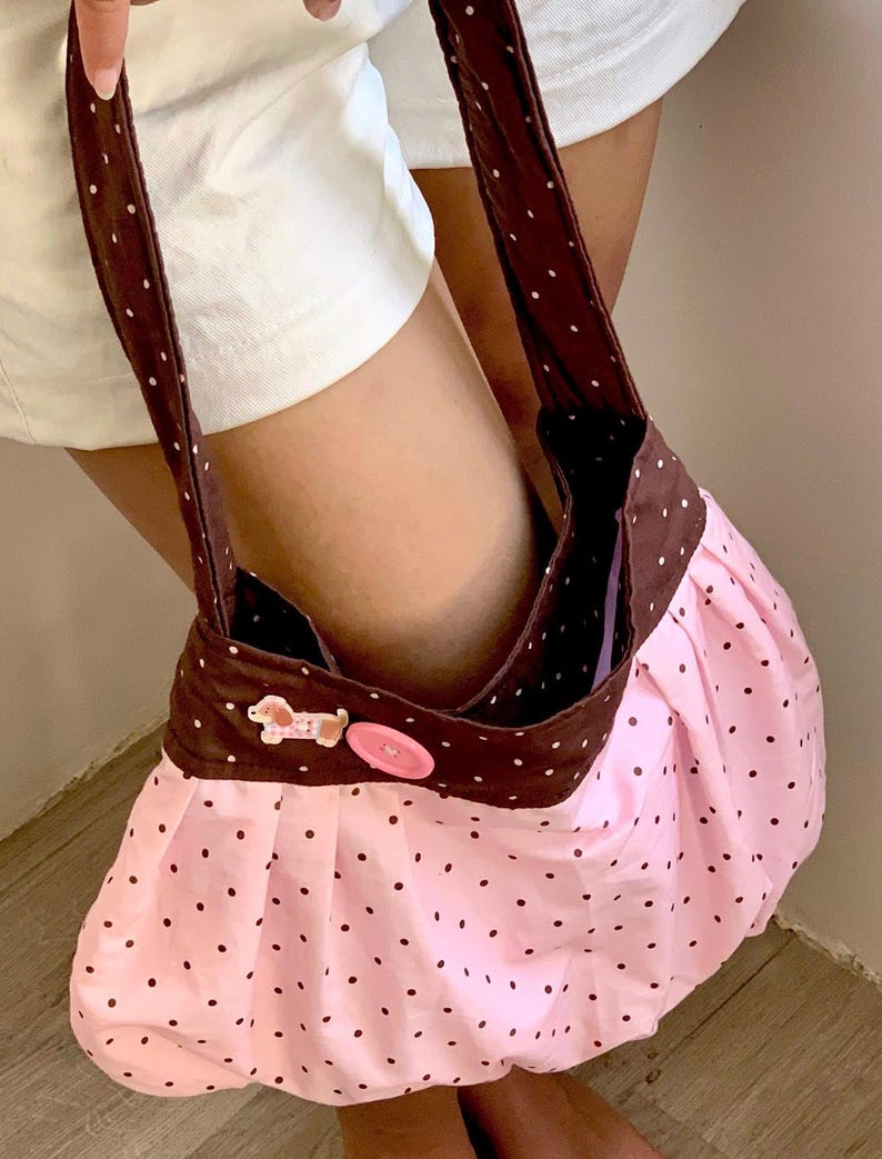 Peut inclure: Un sac &agrave; main rose &agrave; pois avec un dessus et des sangles marron. Le sac est orn&eacute; d'un bouton d&eacute;coratif et d'un petit ornement en forme de chien. Le sac est en tissu et a un design fronc&eacute;.