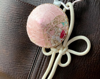 May include: A pink, round fabric charm with a floral design, attached to a white cord and silver-tone metal clasp. The charm is hanging from a brown leather bag.