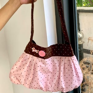 Peut inclure: Un sac &agrave; main rose et marron &agrave; pois, de forme arrondie. Le sac pr&eacute;sente un corps rose &agrave; pois noirs et un dessus marron &agrave; pois blancs. Un bouton rose et un petit &eacute;l&eacute;ment d&eacute;coratif ornent le devant. Le sac a des anses marron.