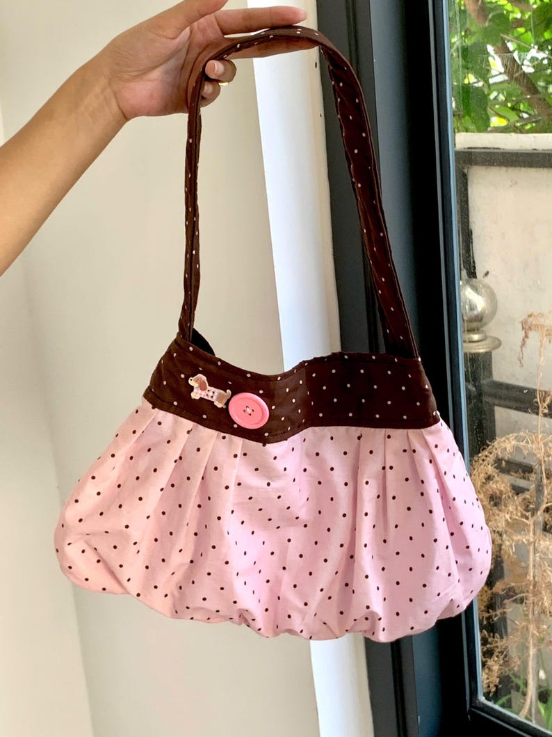Peut inclure: Un sac &agrave; main rose et marron &agrave; pois, de forme arrondie. Le sac pr&eacute;sente un corps rose &agrave; pois noirs et un dessus marron &agrave; pois blancs. Un bouton rose et un petit &eacute;l&eacute;ment d&eacute;coratif ornent le devant. Le sac a des anses marron.