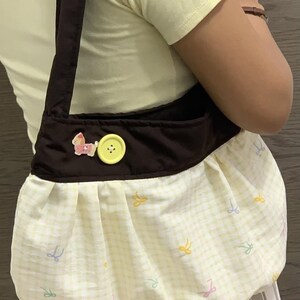 May include: A brown shoulder bag with a yellow and white checkered pattern. The bag features a brown strap, a yellow button, and a pink horse applique. The bag's lower section has a gathered design with pastel bow accents.