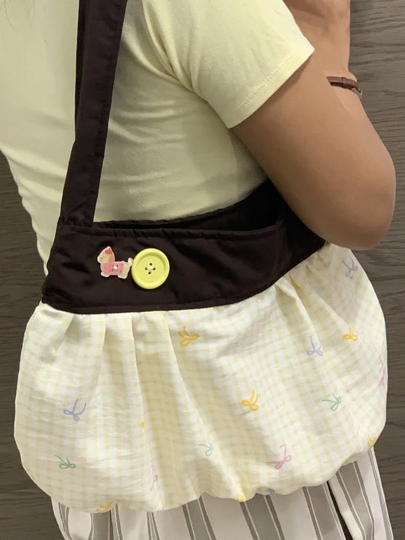 May include: A brown shoulder bag with a yellow and white checkered pattern. The bag features a brown strap, a yellow button, and a pink horse applique. The bag's lower section has a gathered design with pastel bow accents.