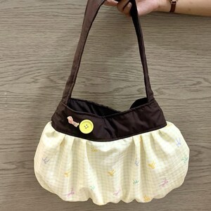 May include: A brown and yellow handbag with a shoulder strap. The bag features a brown top and a yellow gingham skirt with colorful bow accents. A yellow button and pink bow detail the front.
