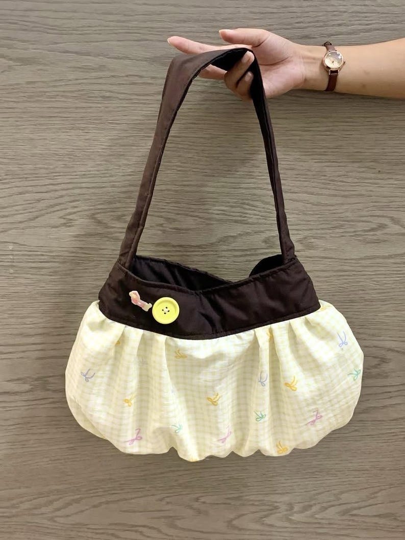 May include: A brown and yellow handbag with a shoulder strap. The bag features a brown top and a yellow gingham skirt with colorful bow accents. A yellow button and pink bow detail the front.